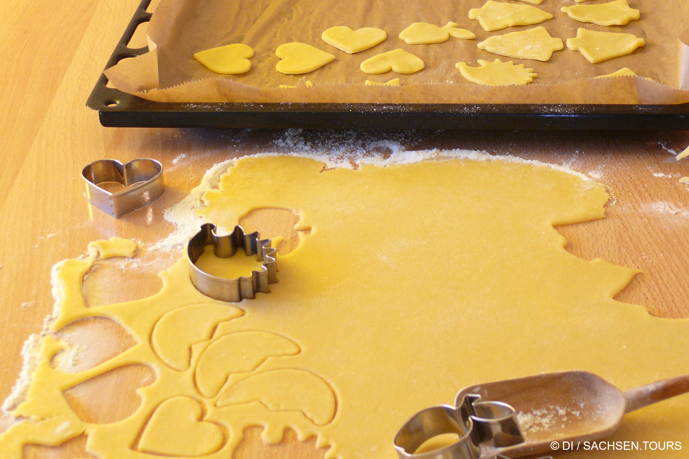 Plätzchenteig aus Omas Grundrezept auf einer bemehlten Fläche ausgerollt und mit Herz- und Sternausstechern ausgestochen. Im Hintergrund steht ein Backblech mit weiteren Plätzchenteig-Kreationen inmitten von verstreutem Mehl.